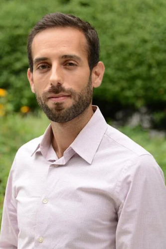 Andrew Albin, a man with facial hair, in a light purple shirt standing in front of yellow flowers