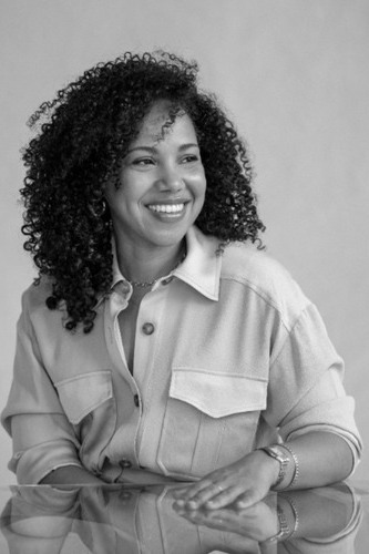 Autumn Womack, a black and white picture of a young dark skinned woman with curly hair smiling, sitting at a reflective table