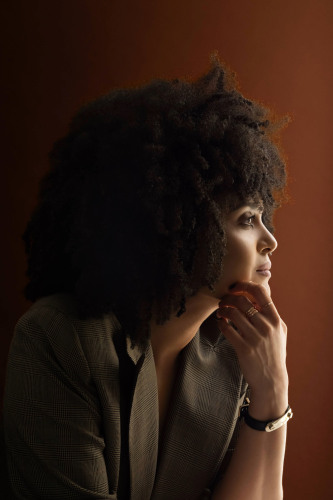 Poet Diannely Antigua, (photo by Beowulf Sheehan), a dark skinned woman with dark curly hair and an nasal piercing in profile facing left with her hand to her chin