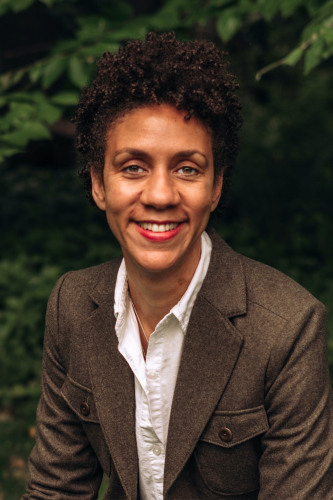 Ebony Flowers- photo by Cate Barry. A dark skinned woman with short curly hair in a brown jacket and white shirt in from of a green leafy background