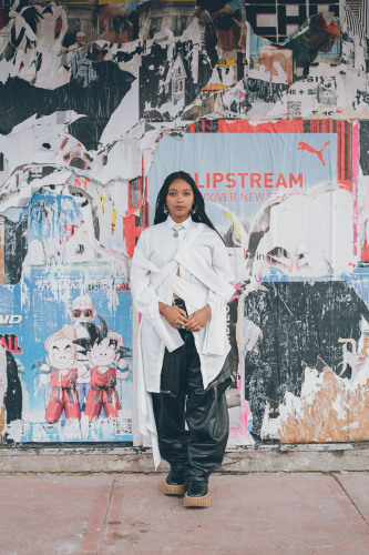 Safia Elhillo, a young woman in a white top standing in front of a large street mural with collaged posters