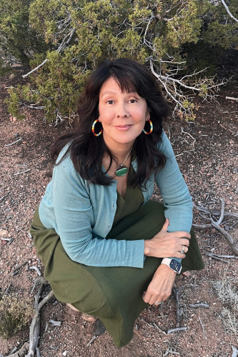 Deborah Taffa- A native american woman in a green dress and teal sweater, and large hoop earrings on a brick squatting down in front of an evergreen