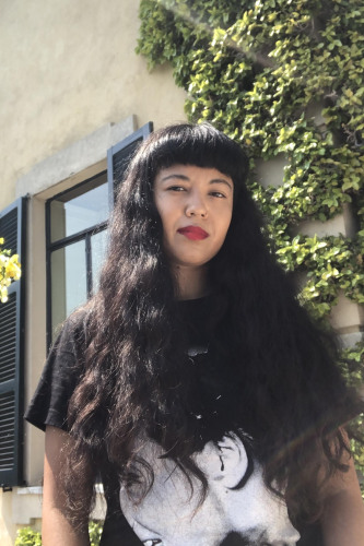 Jackie Wang, a woman with long dark hair and red lips, outside in front of a building with a pot of yellow flowers to her right