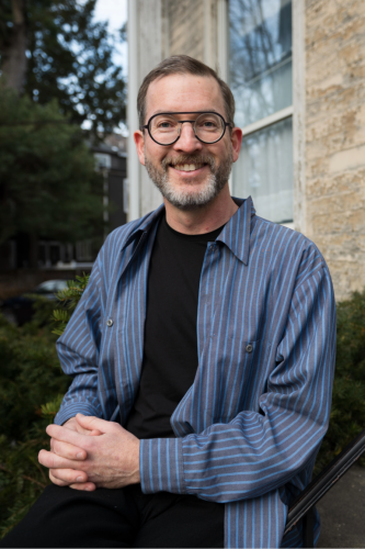 Jonathan Schroeder, photo by Kevin Grady/Harvard Radcliffe Institute. A man with glasses and facial hair in an un buttoned Blue striped dress shirt with Black Tshirt underneith smiles nea a handrail outside